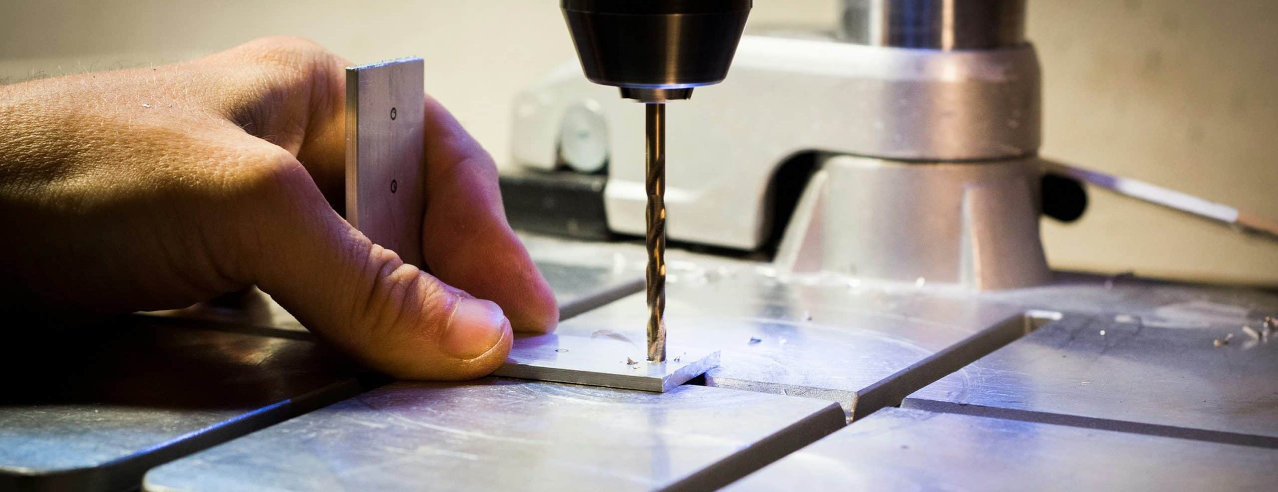 A close-up shot of an industrial drill boring into a piece of metal, with a hand holding the metal firmly in place.