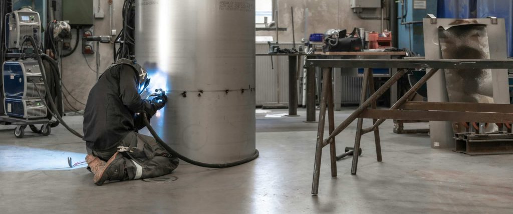 A man wearing a welding mask and protective safety gear welds a metal tube, with bright sparks flying from the torch. Photo by Jimmy Nilsson-Masth.
